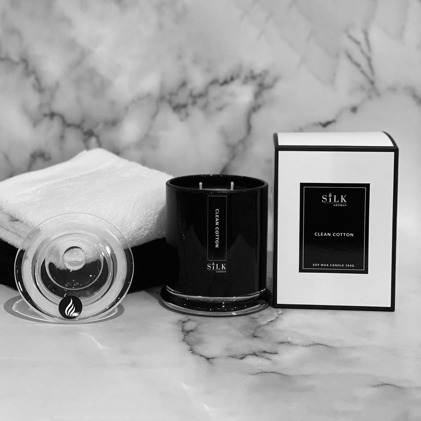 A black glossy candle jar with clear glass apothecary lid, next to a luxury white box with a black label and black trim