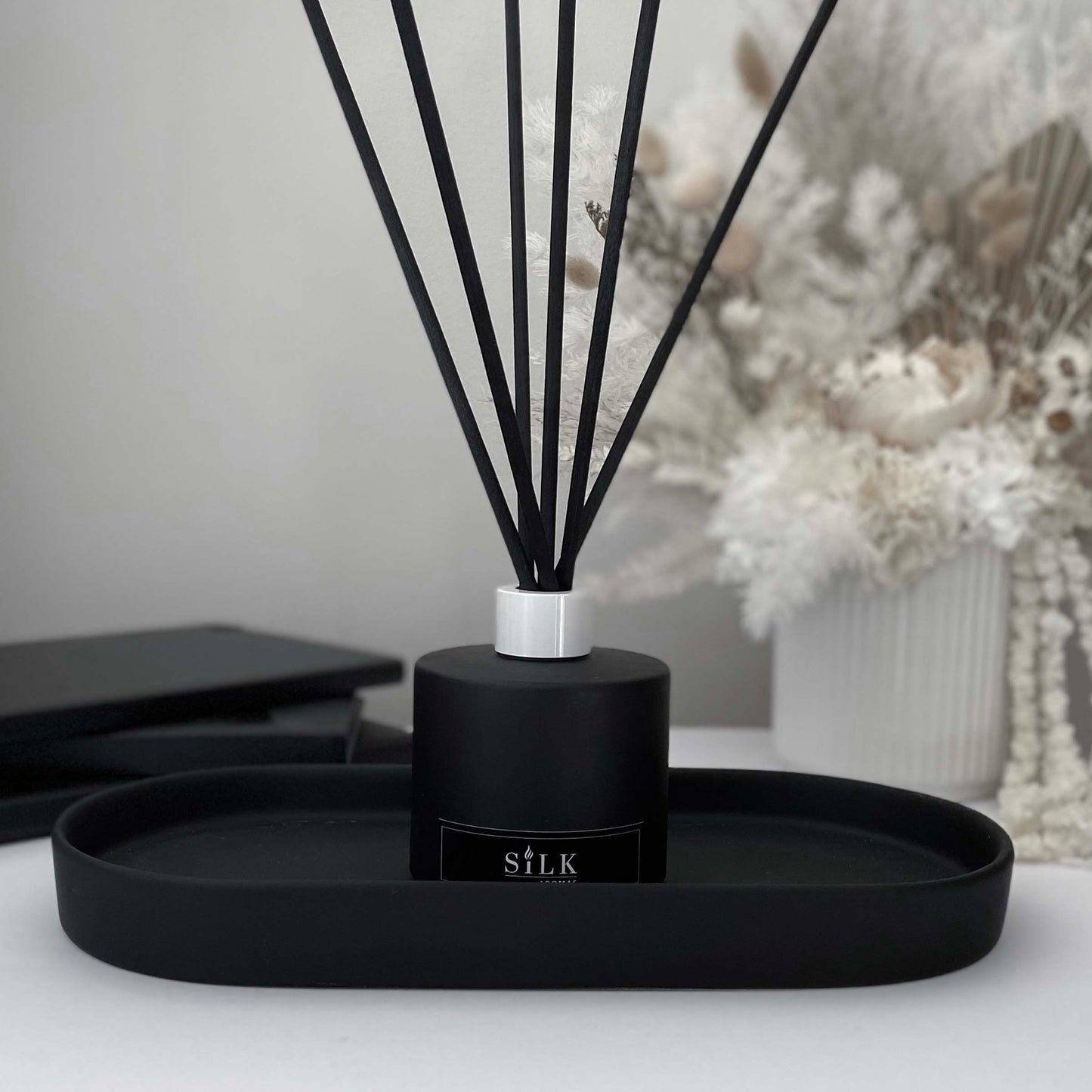 A black diffuser bottle with a white lid and black diffuser reeds on a black tray, with a white floral arrangement behind it