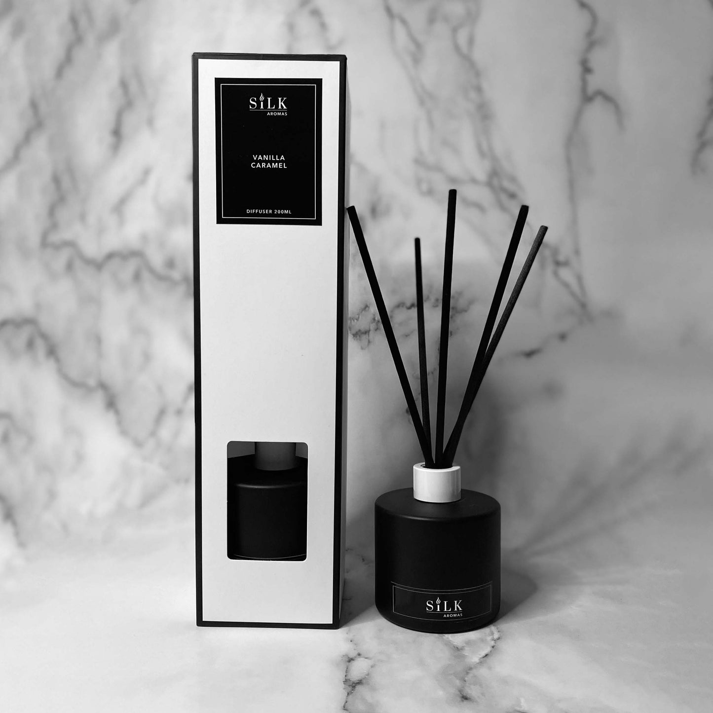 A black diffuser bottle with a white lid and black diffuser reeds, next to the same in a white box with a black label and black trim