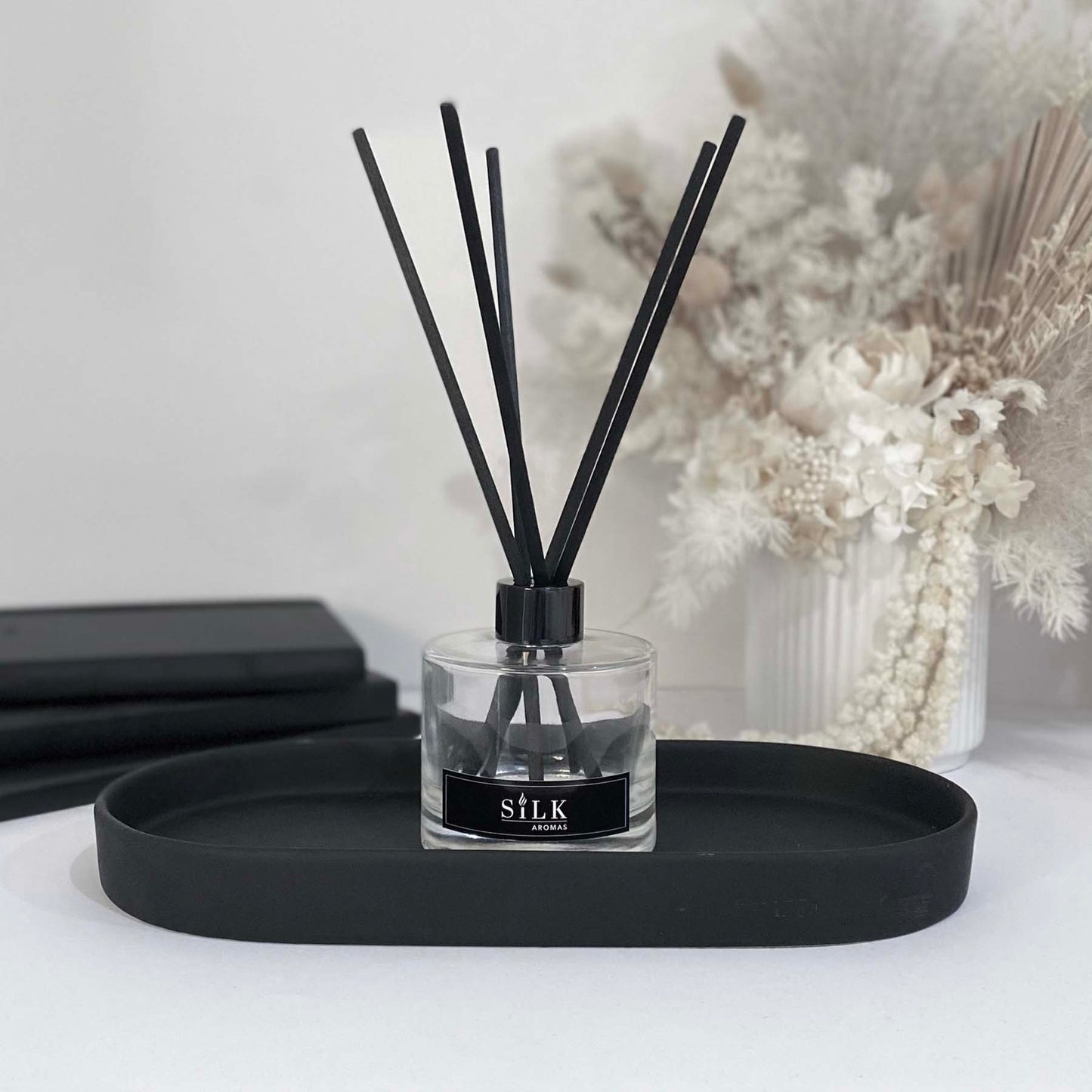A clear diffuser bottle with a black lid and black diffuser reeds on a black tray, with a white floral arrangement behind it