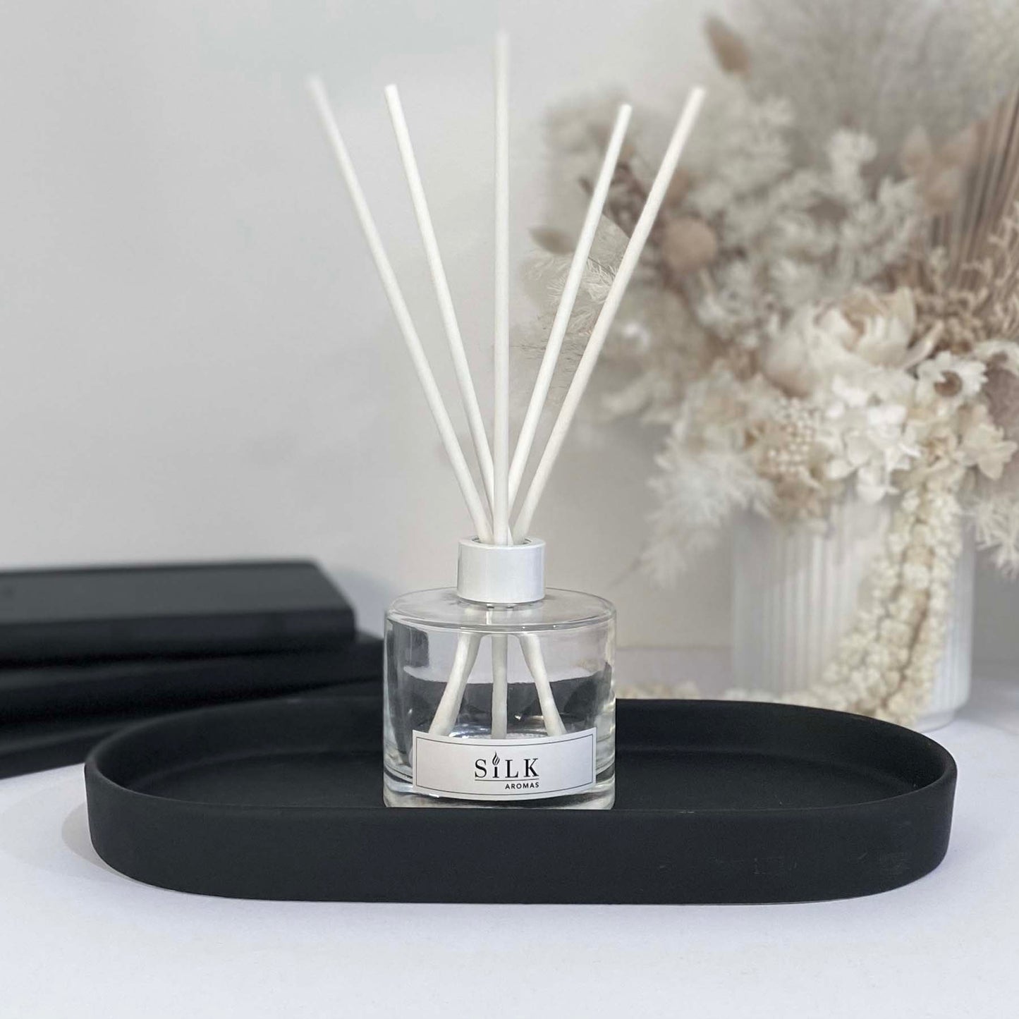 A clear diffuser bottle with a white lid and white diffuser reeds on a black tray, with a white floral arrangement behind it