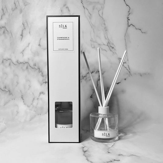 A clear diffuser bottle with a white lid and white diffuser reeds, next to the same in a white box with a white label and black trim