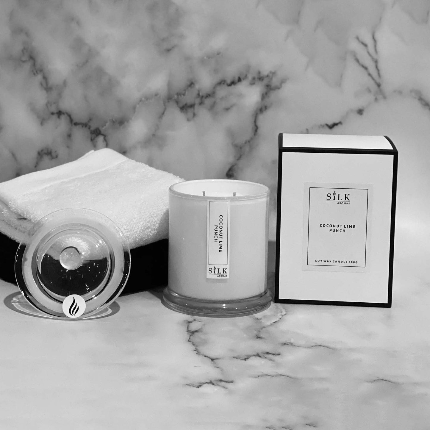 A white glossy candle jar with clear glass apothecary lid, next to a luxury white box with a white label and black trim
