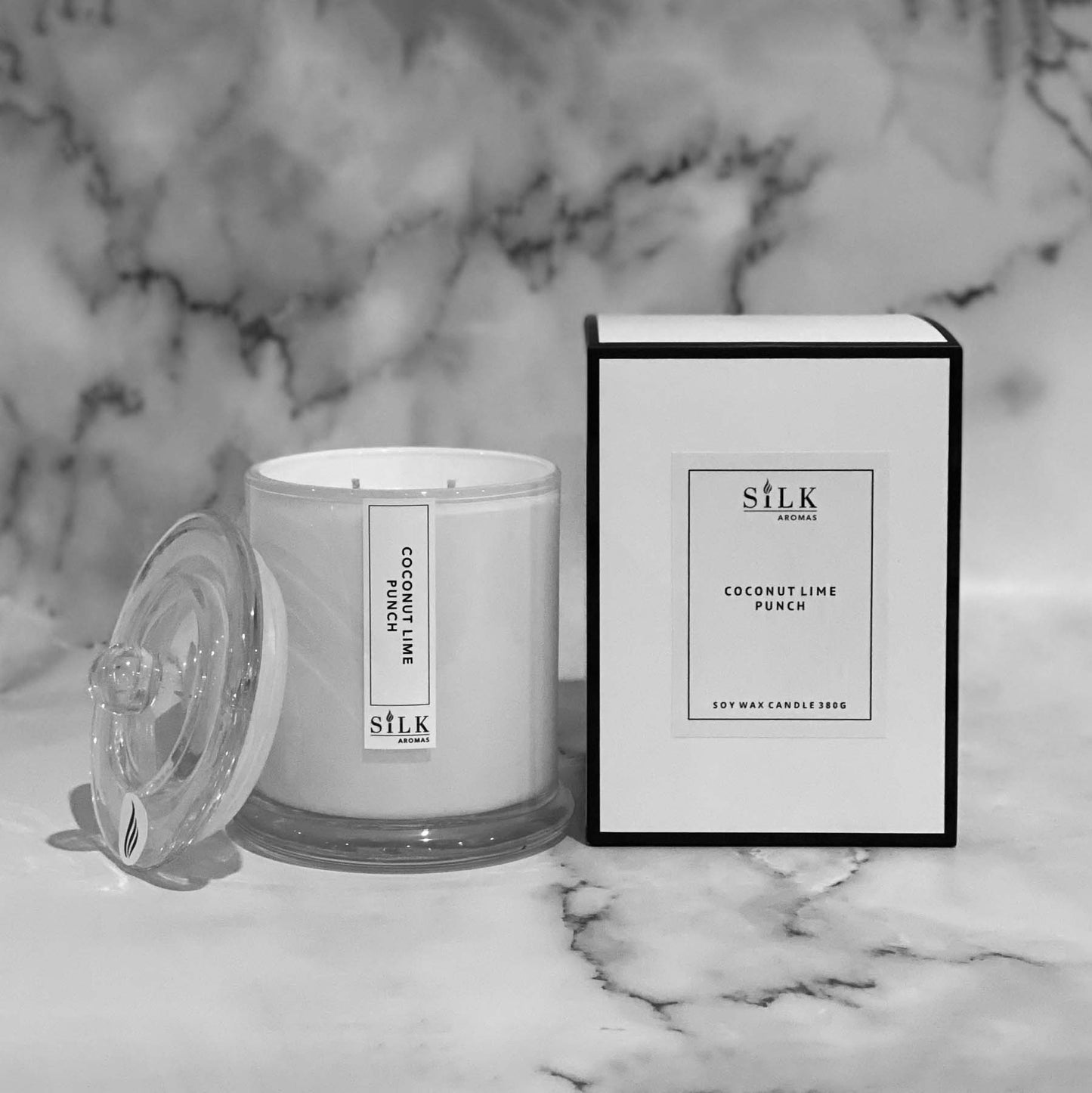 A white glossy candle jar with clear glass apothecary lid, next to a luxury white box with a white label and black trim