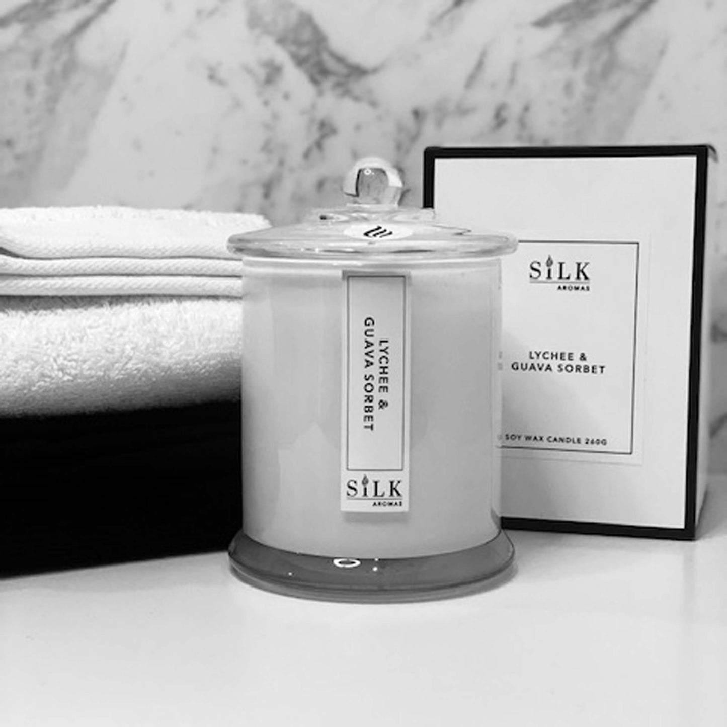 A white glossy candle jar with clear glass apothecary lid, next to a luxury white box with a white label and black trim
