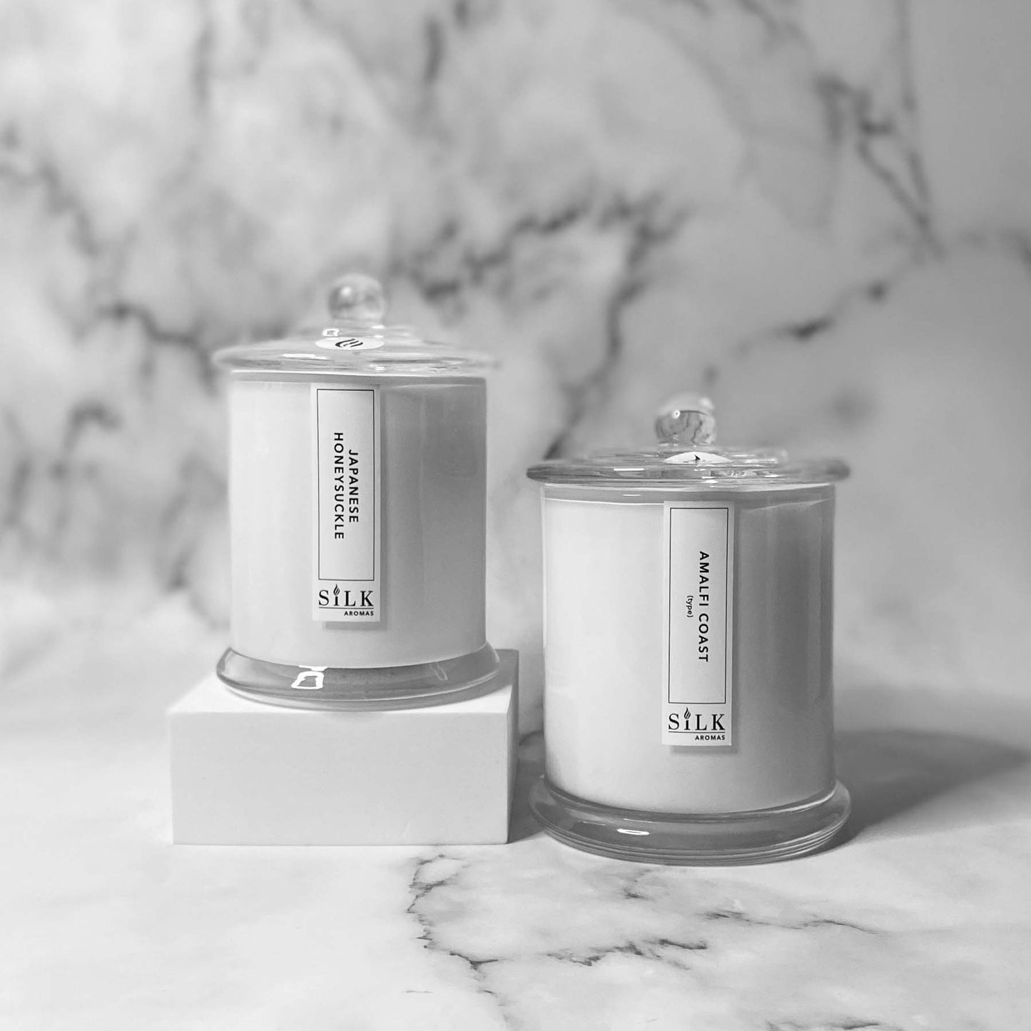 Two white candles, one smaller and one larger, in glossy white jars with clear glass apothecary lids
