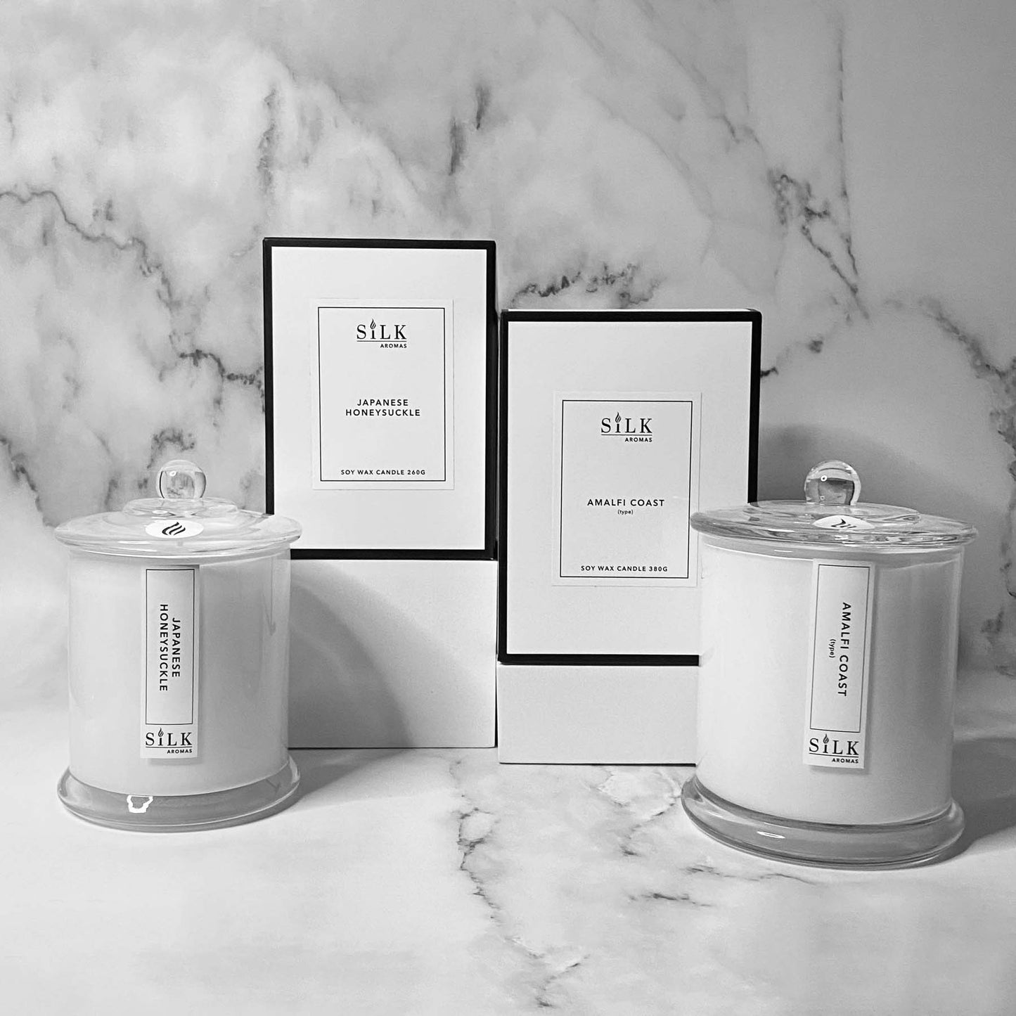 White glossy candles in two sizes with clear glass apothecary lids, next to luxury white boxes with a white label and black trim
