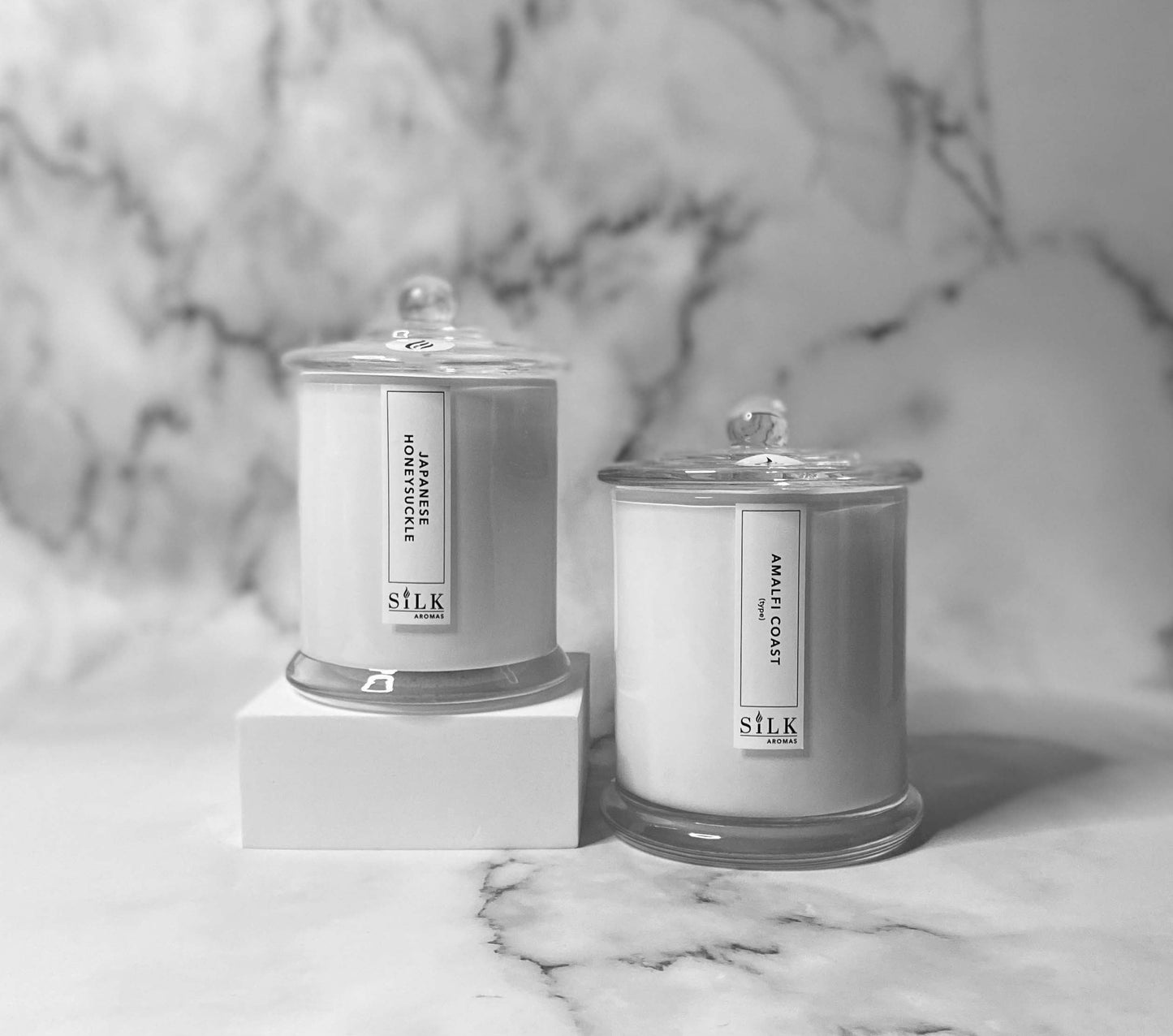 Two white candles, one smaller and one larger, in glossy white jars with clear glass apothecary lids