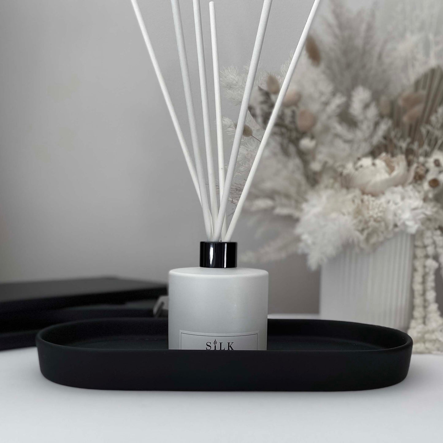A white diffuser bottle with a black lid and white diffuser reeds on a black tray, with a white floral arrangement behind it