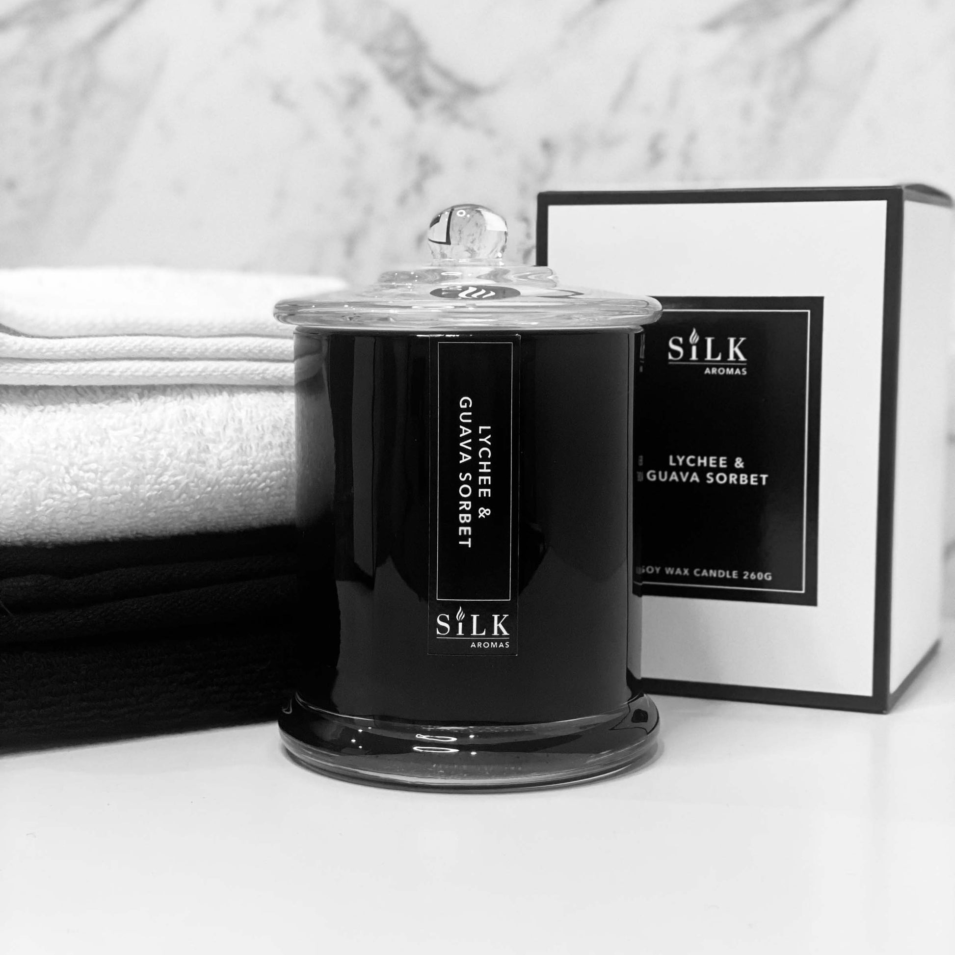 A black glossy candle jar with clear glass apothecary lid, next to a luxury white box with a black label and black trim