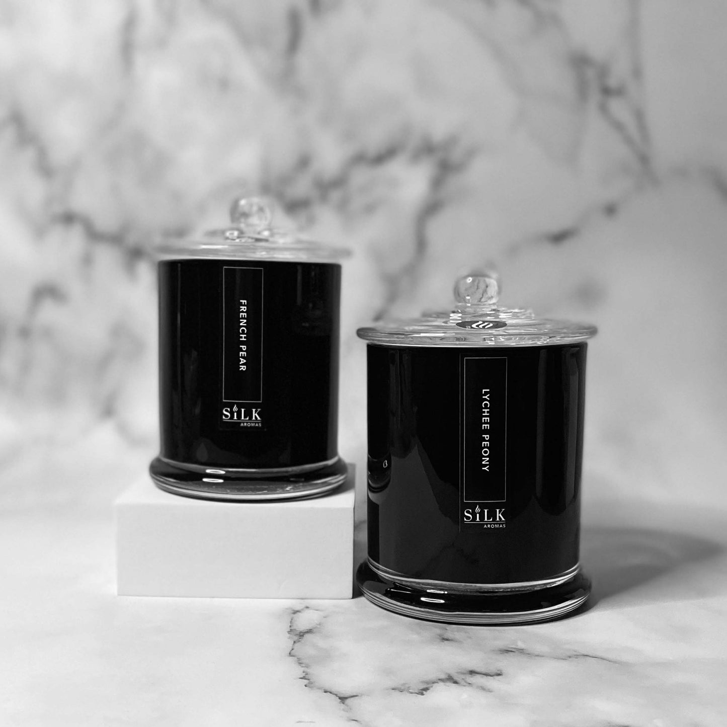 Two candles, one smaller and one larger, in glossy black jars with clear glass apothecary lids